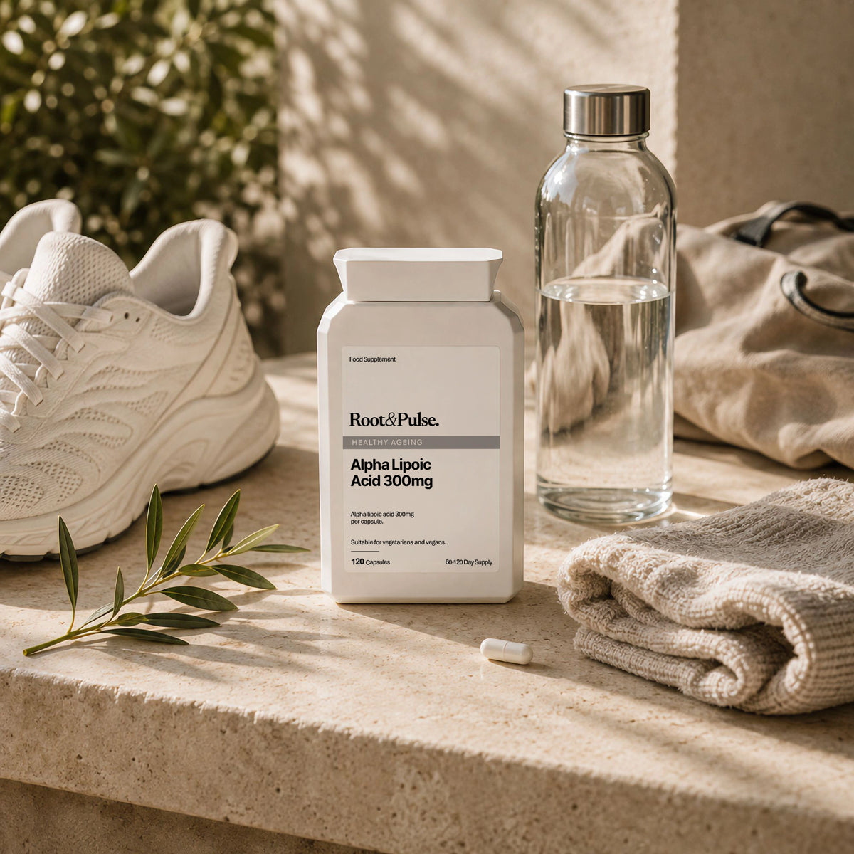 A white bottle of ROOT & PULSE Alpha Lipoic Acid 300mg capsules placed on a stone ledge outdoors, surrounded by a white sneaker, a clear water bottle, a folded beige towel, and a small olive sprig.
