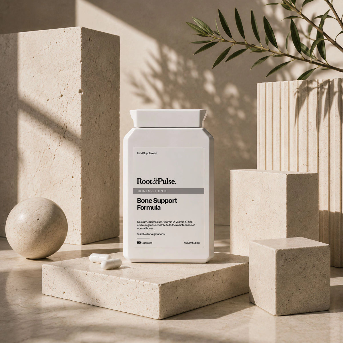 A white bottle of Root & Pulse Bone Support Formula stands on a beige stone pedestal in a styled studio scene with geometric blocks, a small capsule, and soft natural shadows.