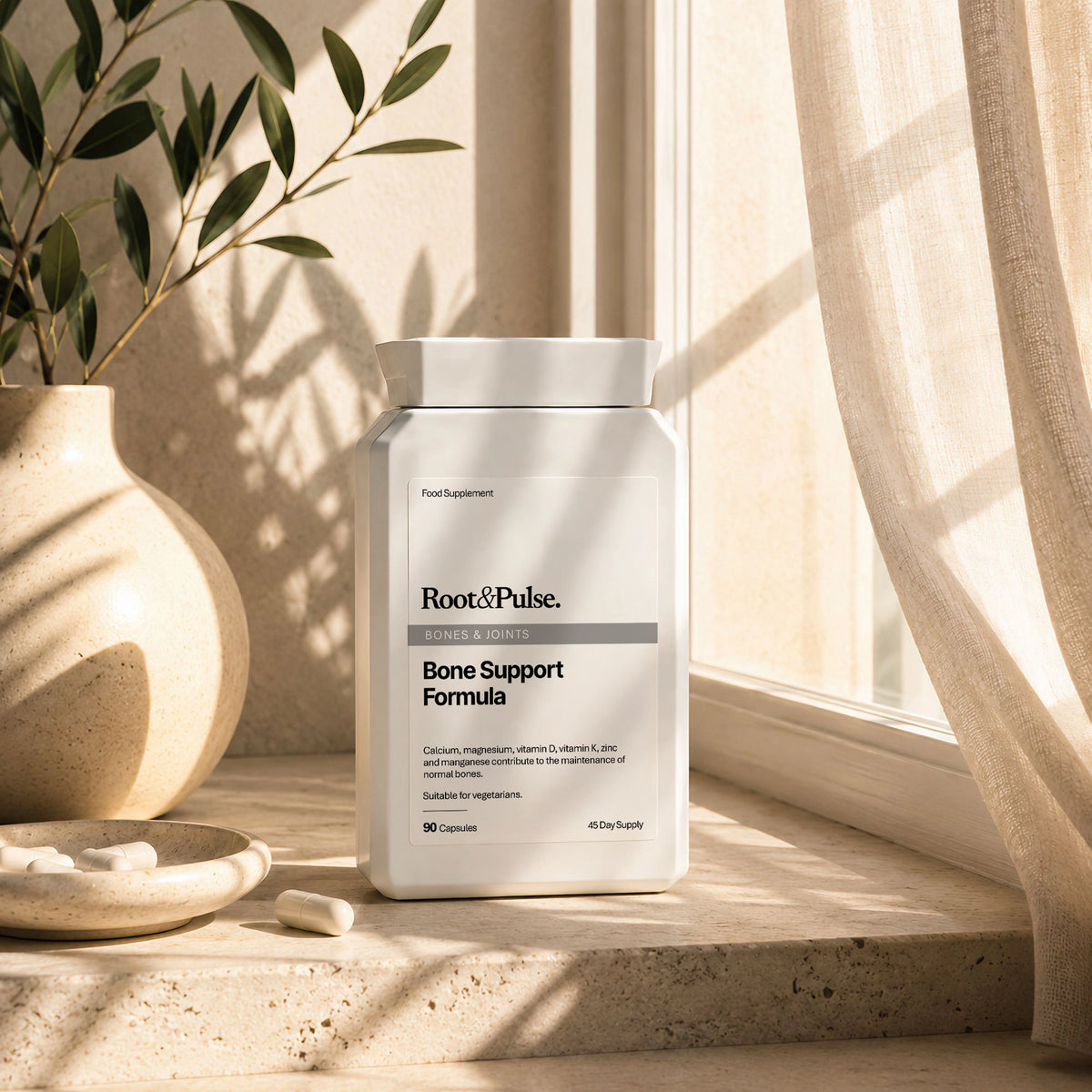 A white supplement bottle labeled Root & Pulse Bone Support Formula sits on a sunlit windowsill with warm natural light, a beige ceramic vase with greenery, and sheer curtains in the background.