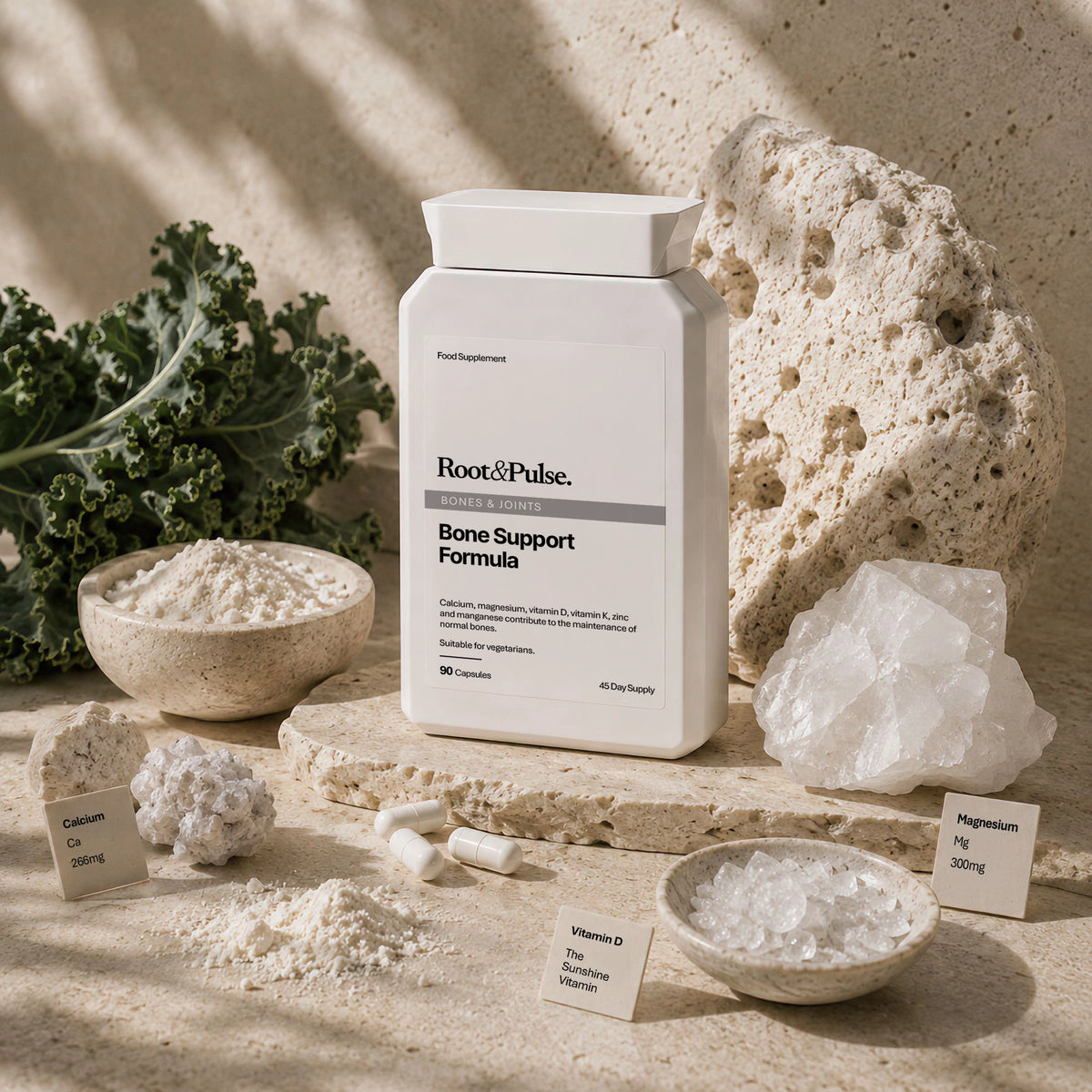 A white supplement bottle labeled Root & Pulse Bone Support Formula stands on a stone slab surrounded by calcium, magnesium, vitamin D powder, kale, and rock props in a warm, sandy studio setting.