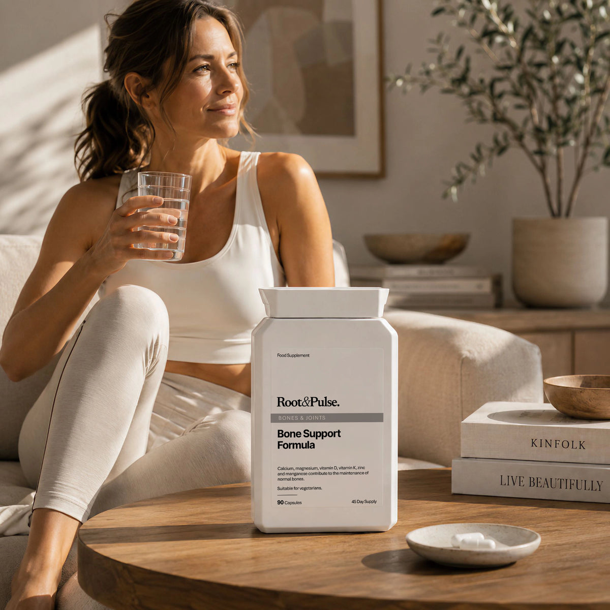 A woman in a white tank top and leggings sits on a beige sofa holding a glass of water, with a white container of Root of Pulse Bone Support Formula placed on a wooden table in front of her in a bright, modern living room.