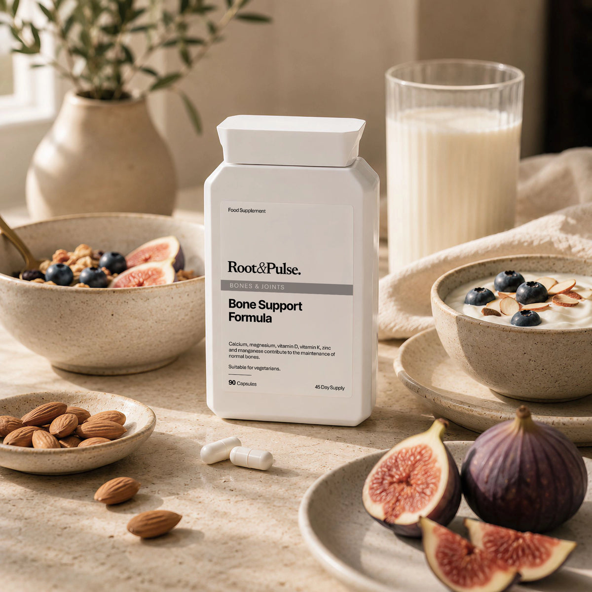 A bottle of Root & Pulse Bone Support Formula stands on a tabletop surrounded by bowls of yogurt with blueberries and figs, almonds, and a glass of milk in a warm, softly lit kitchen setting.