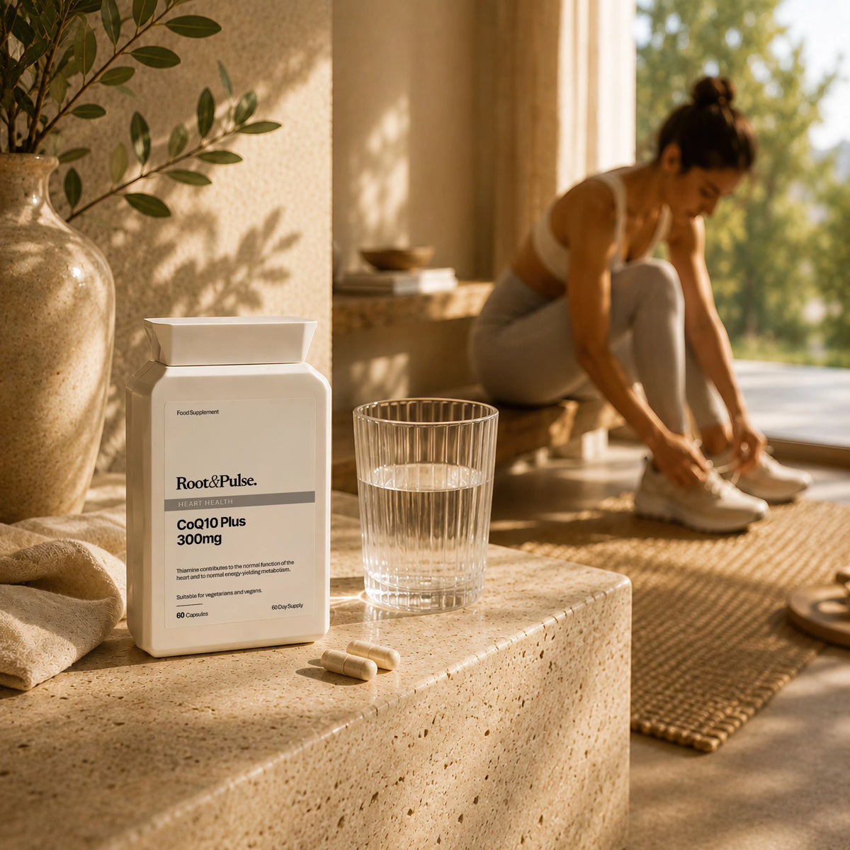A white supplement bottle labeled Root & Pulse Co Q10 Plus 300mg sits on a stone ledge beside a glass of water and a few capsules, with a woman sitting in the background by a window.