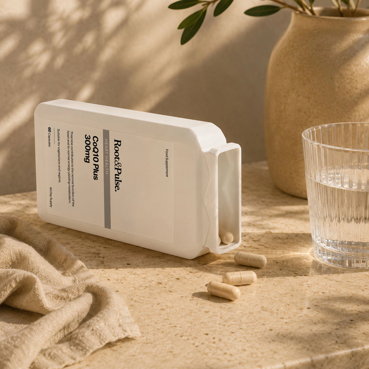 A white bottle of Rose & Pulse Co-Q10 Plus capsules lies on its side on a beige stone surface, with several capsules spilled out. A glass of water, a beige vase, and a beige cloth are nearby in a warm, sunlit setting.