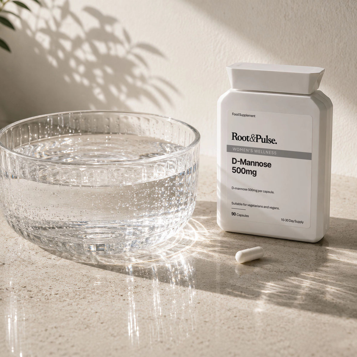 A white bottle of Root & Pulse D-Mannose 500mg dietary supplement stands on a light stone surface beside a clear glass bowl of water, with a single capsule lying in front. The scene is lit by warm natural sunlight with leaf shadows cast across the background wall.