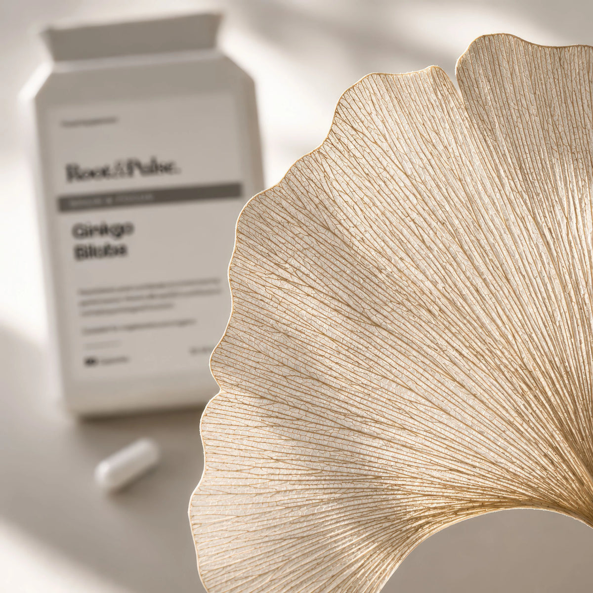 A close-up of a gold ginkgo leaf-shaped object in sharp focus, with a white Root & Pulse Ginkgo Blume supplement bottle and capsule blurred in the background on a light surface.