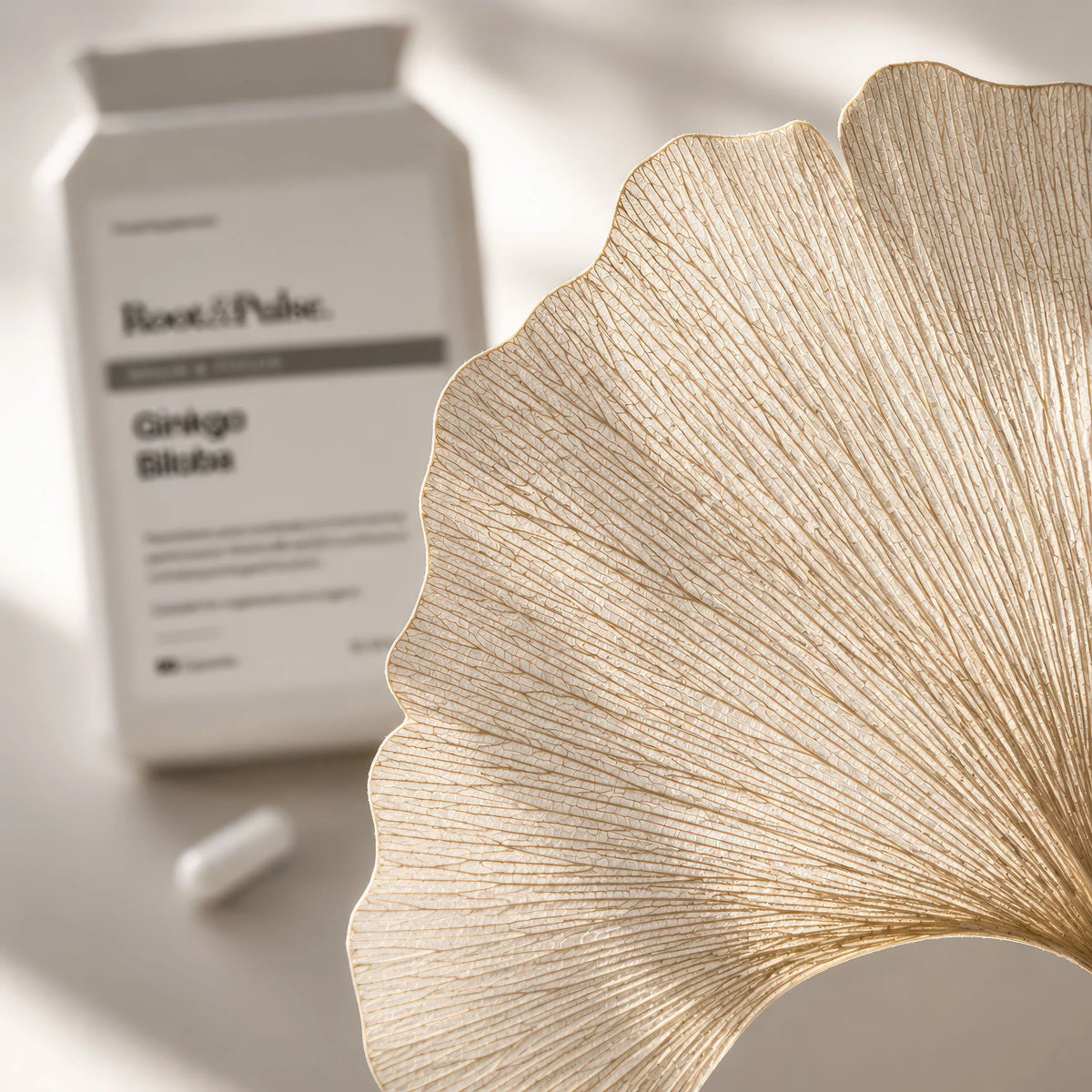 A close-up of a gold ginkgo leaf-shaped object in sharp focus, with a white Root & Pulse Ginkgo Blume supplement bottle and capsule blurred in the background on a light surface.