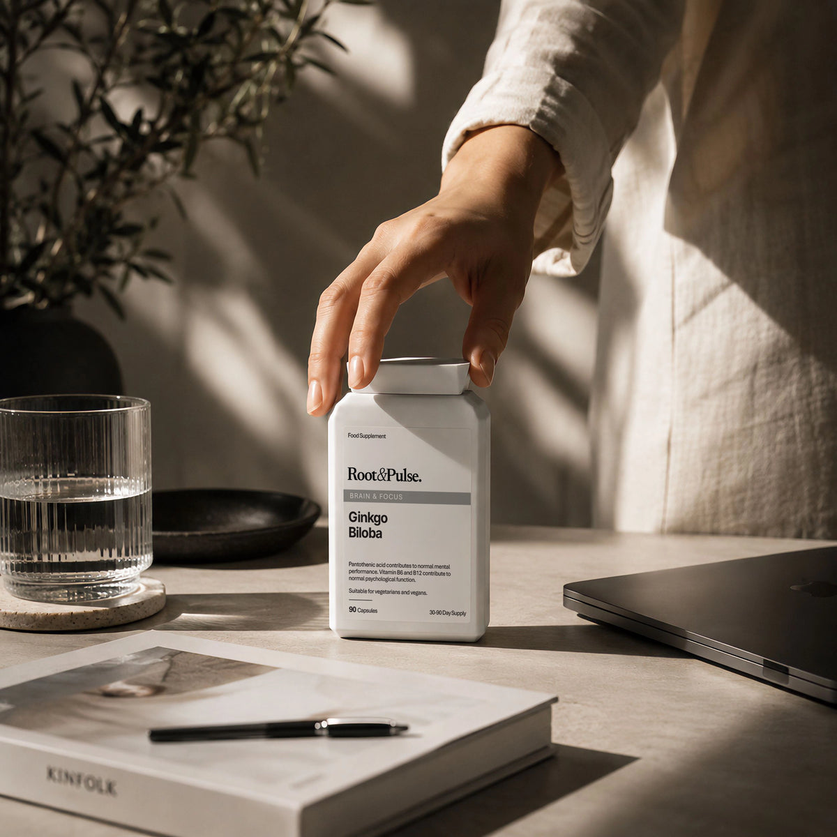 A person's hand holding a white bottle of Root of Pulse Ginkgo Biloba Supplement on a light gray desk with warm sunlight and shadows, surrounded by a glass of water, a book, and a laptop.