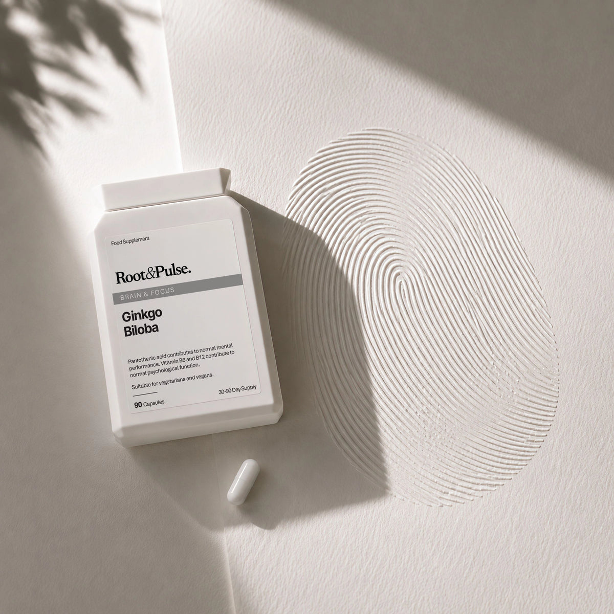 A white bottle of Root & Pulse Ginkgo Biloba capsules with a single capsule beside it, styled on a textured white surface with a large fingerprint pattern and leaf shadows.