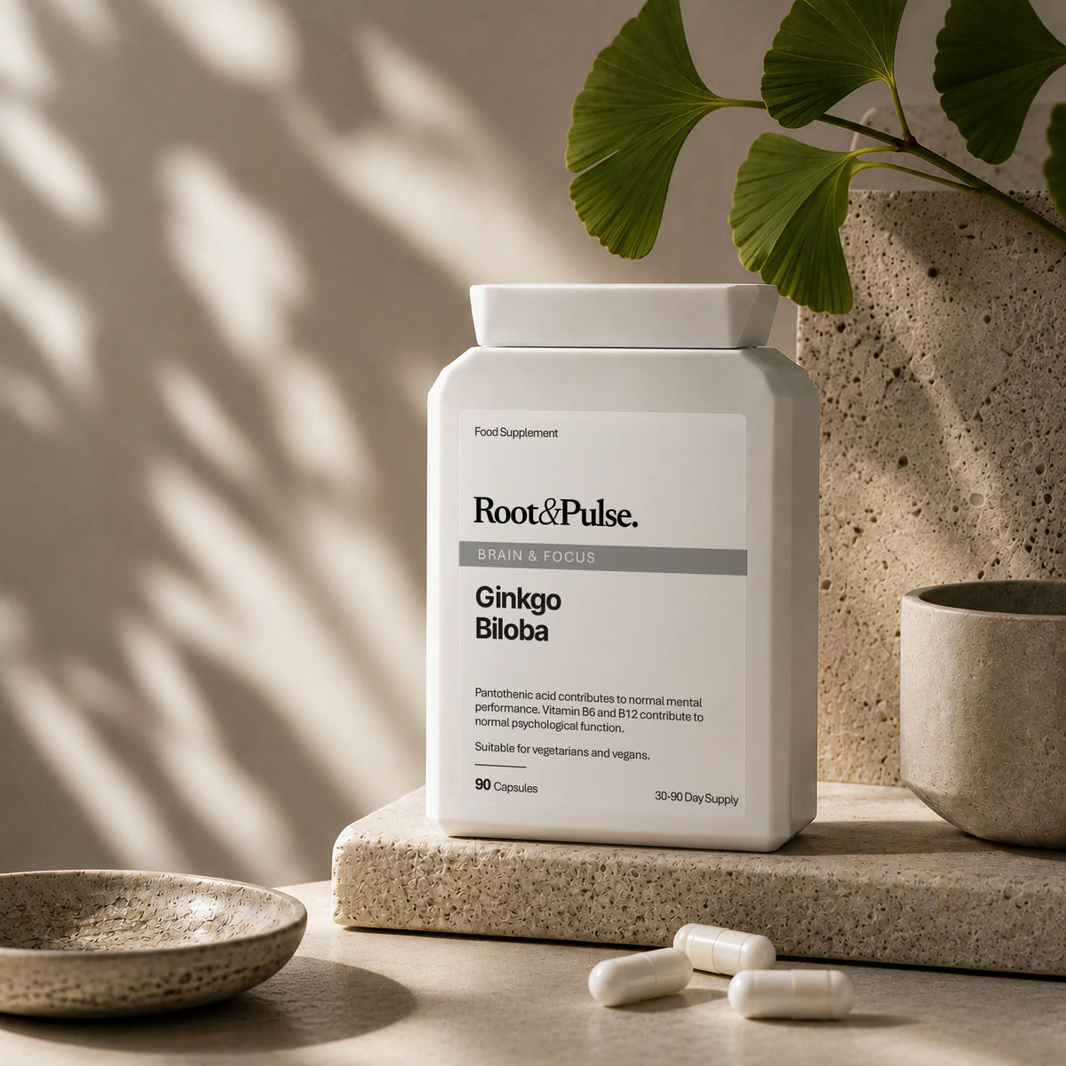 A white bottle of Root & Pulse Ginkgo Biloba dietary supplement capsules, styled on a stone slab with ginkgo leaves and ceramic dishes in a warm, sunlit setting.