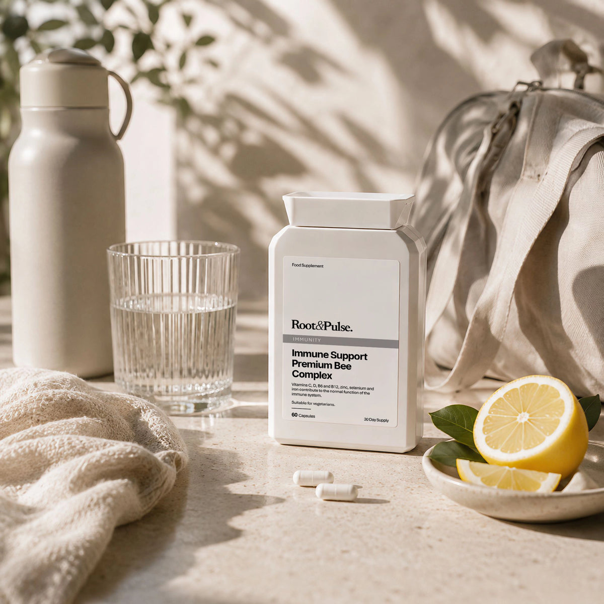 A white bottle of Root of Pulse Immune Support Premium Bee Complex stands on a light stone surface with lemon slices, a glass of water, a beige bottle, and a beige bag in a sunlit setting.