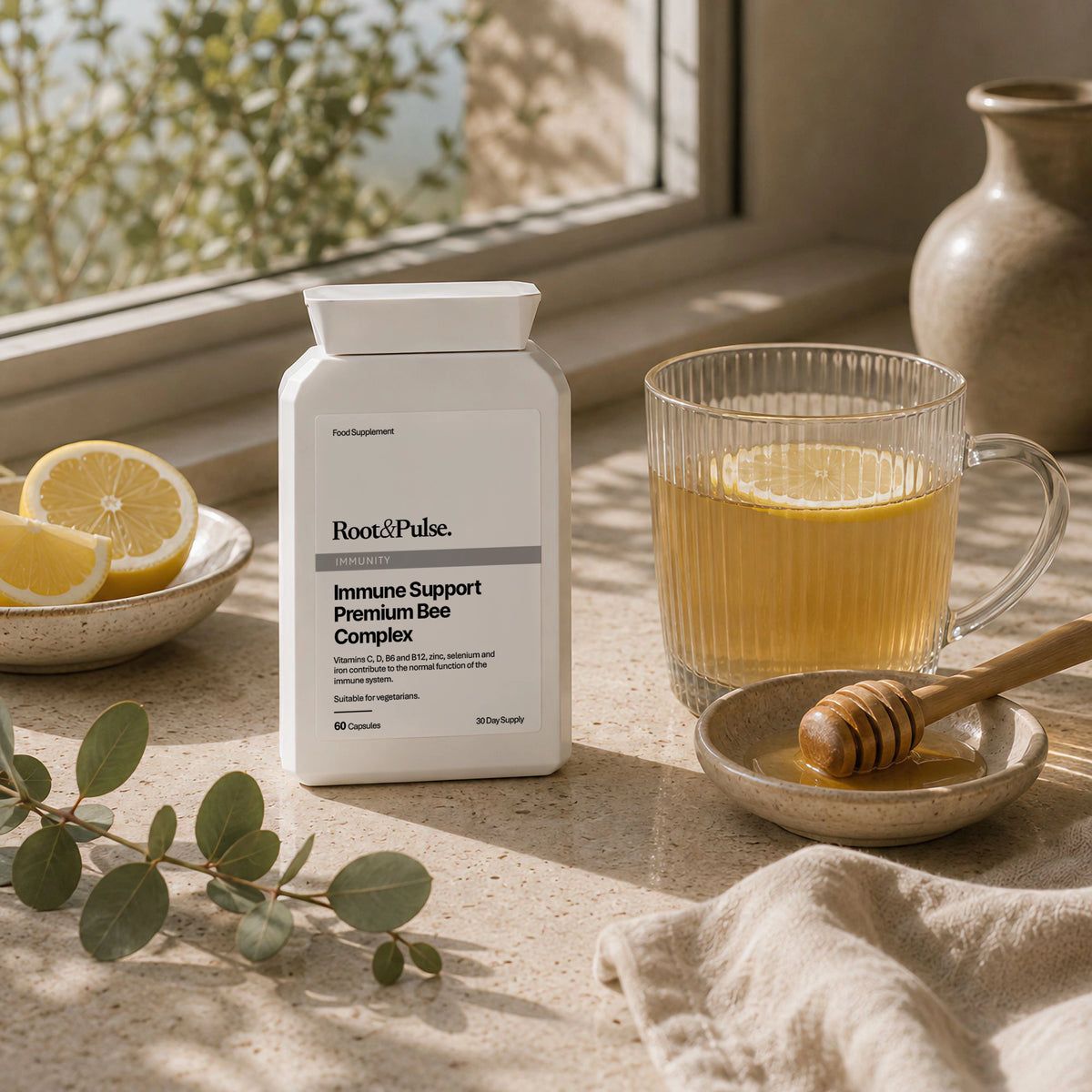 A white bottle of Root&Pulse Immune Support Premium Bee Complex supplement placed on a stone surface beside a glass of honey tea, lemon slices, a honey dipper, and eucalyptus leaves in warm natural light.