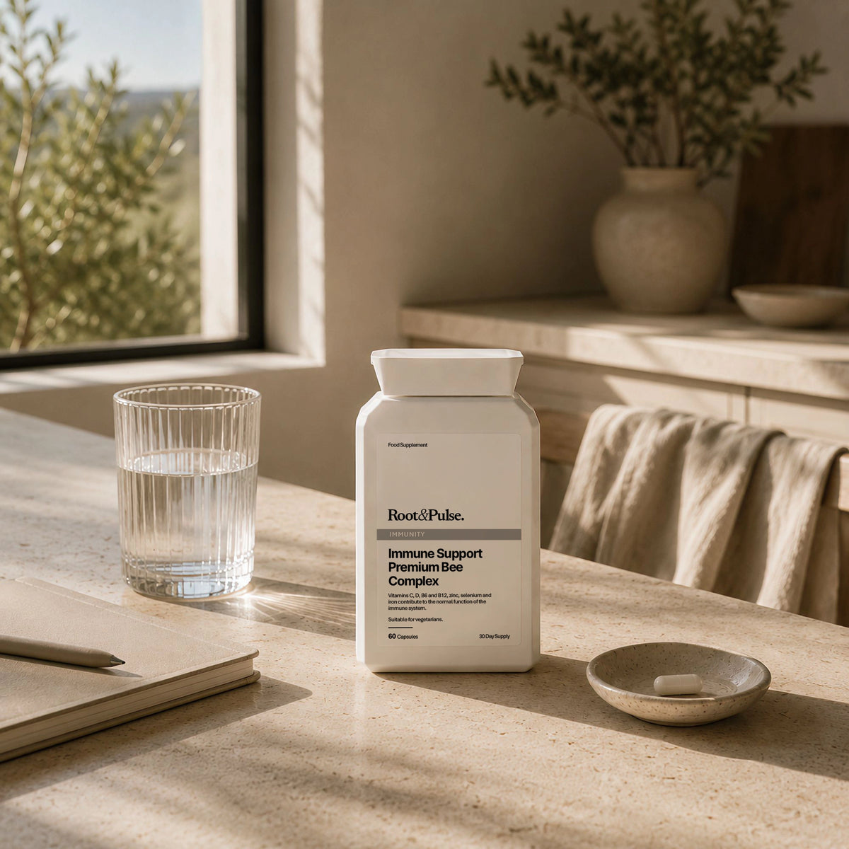 A white bottle of Root of Pulse Immune Support Premium Bee Complex sits on a sunlit stone countertop beside a glass of water, a notebook, and a small dish with a tablet, in a warm, minimalist interior setting.