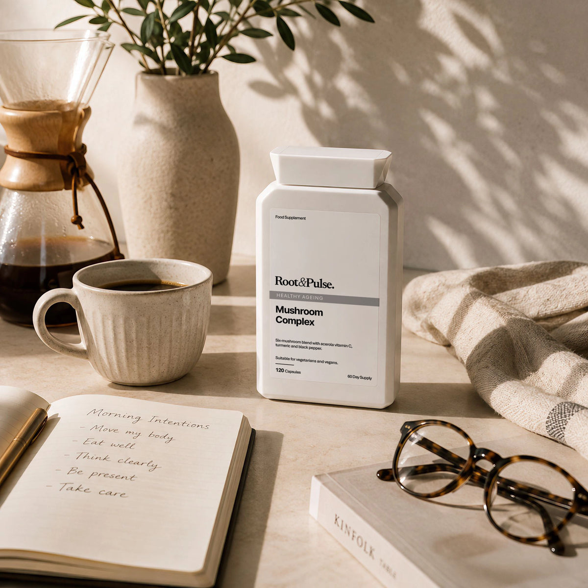 A white bottle of Root & Pulse Mushroom Complex supplement placed on a styled desk with a coffee mug, coffee maker, open notebook, eyeglasses, and a folded blanket in warm natural light.