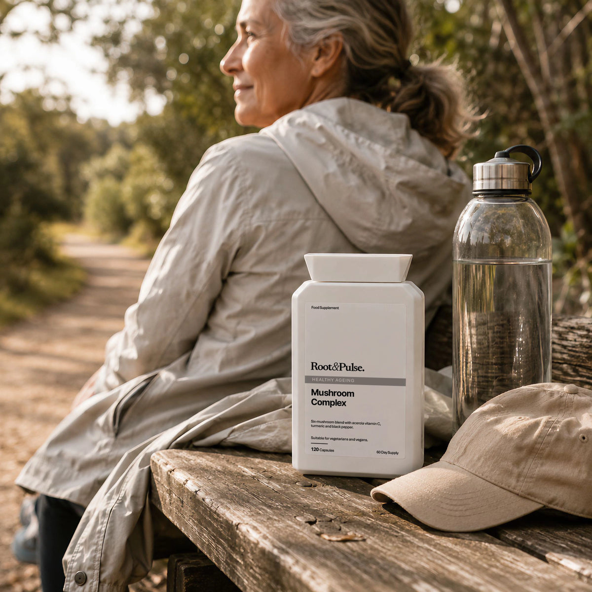 A woman sits on a wooden bench outdoors by a stream, with a white container of Root and Pulse Mushroom Blend Supplement in the foreground and a clear water bottle beside it.