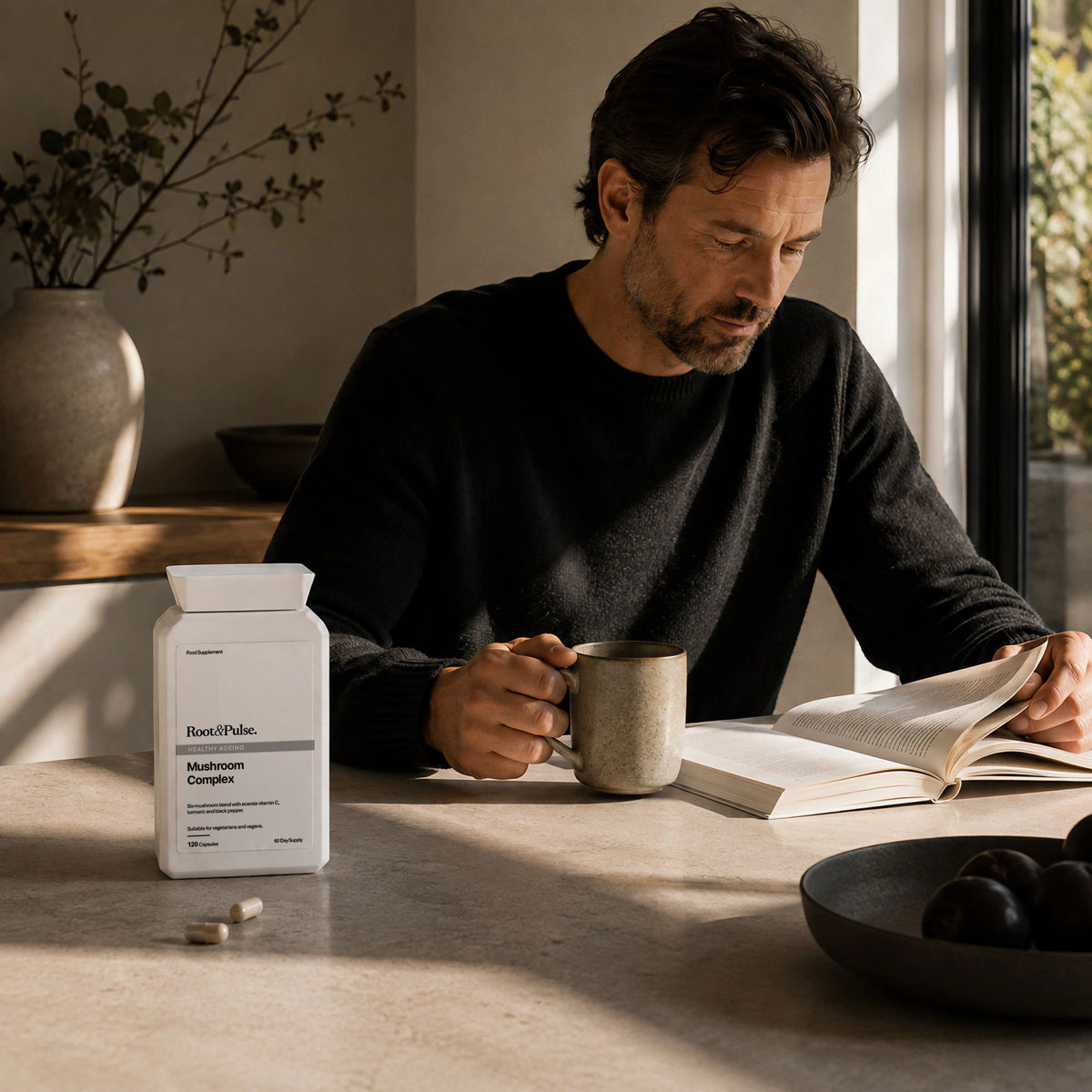 A man in a black sweater sits at a table by a window, reading a book and holding a mug, with a white bottle of Root & Pulse 6 Mushroom Blend Supplement capsules on the table beside him.