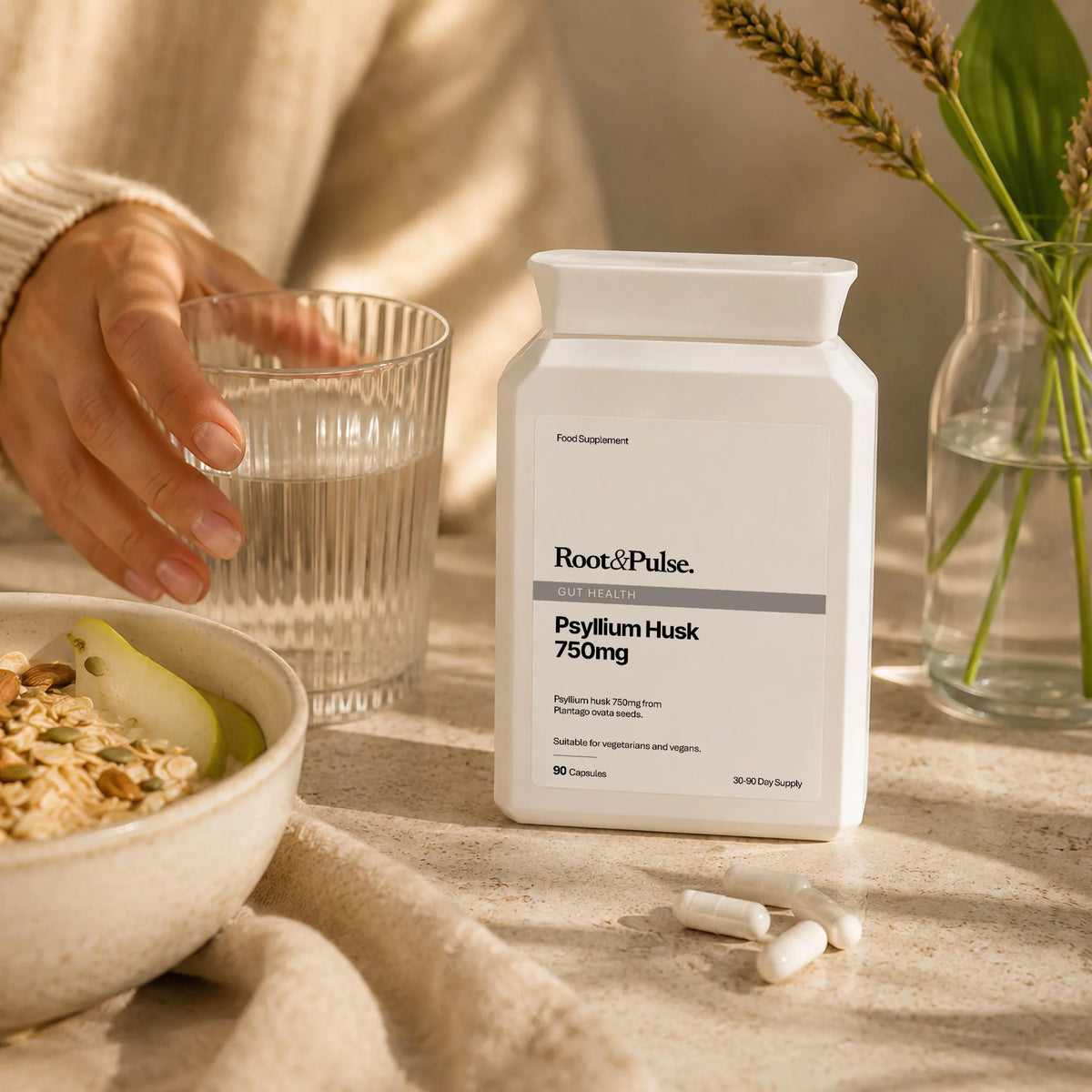 A white bottle of Root & Pulse Psyllium Husk 750mg dietary supplement stands on a beige tabletop, with a hand holding a glass of water nearby and capsules scattered in front. A bowl of oatmeal and a vase with wheat are softly blurred in the background.