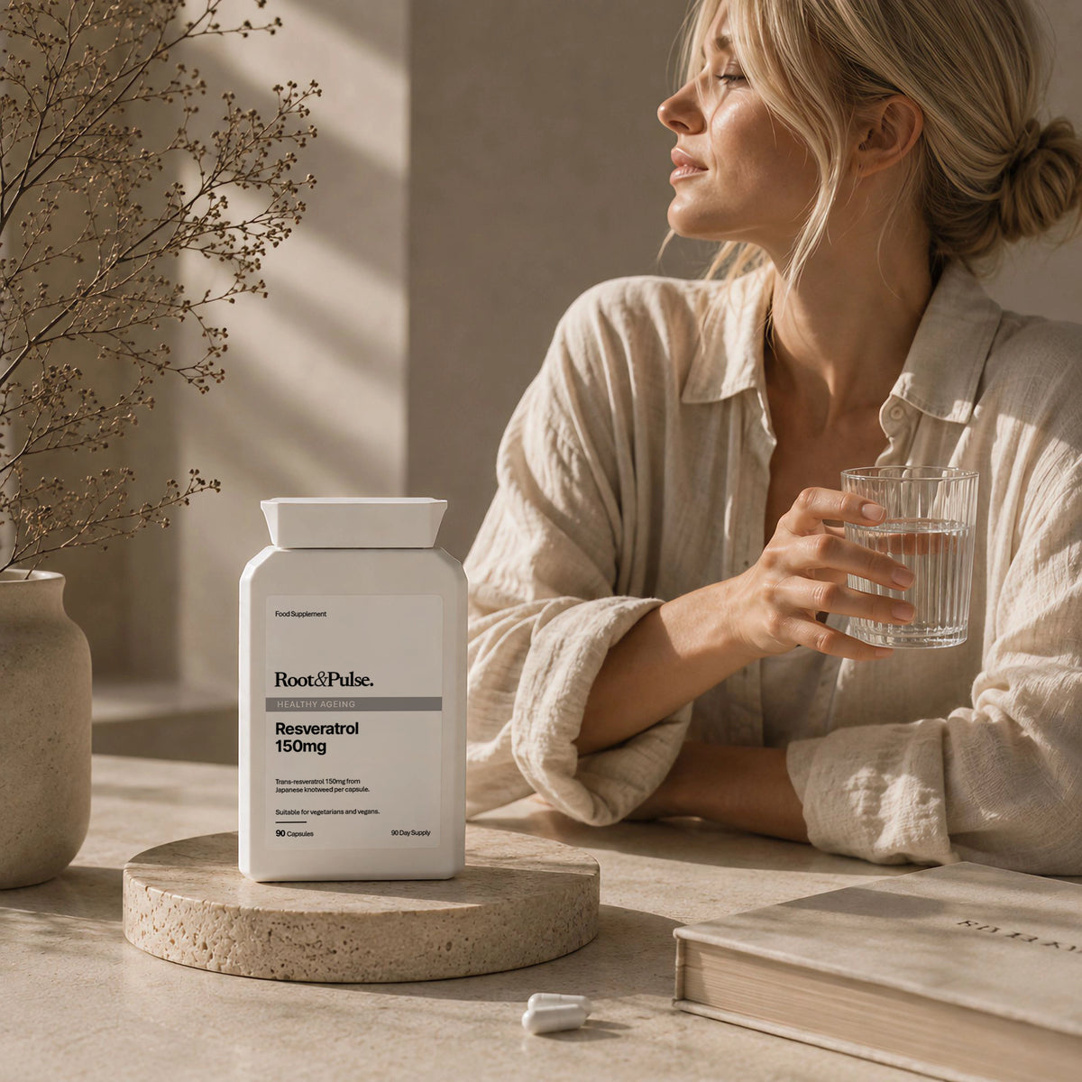 A woman in a beige linen shirt sits at a table holding a glass of water, with a white bottle of Root & Pulse Resveratrol 150mg placed on a stone coaster in front of her.