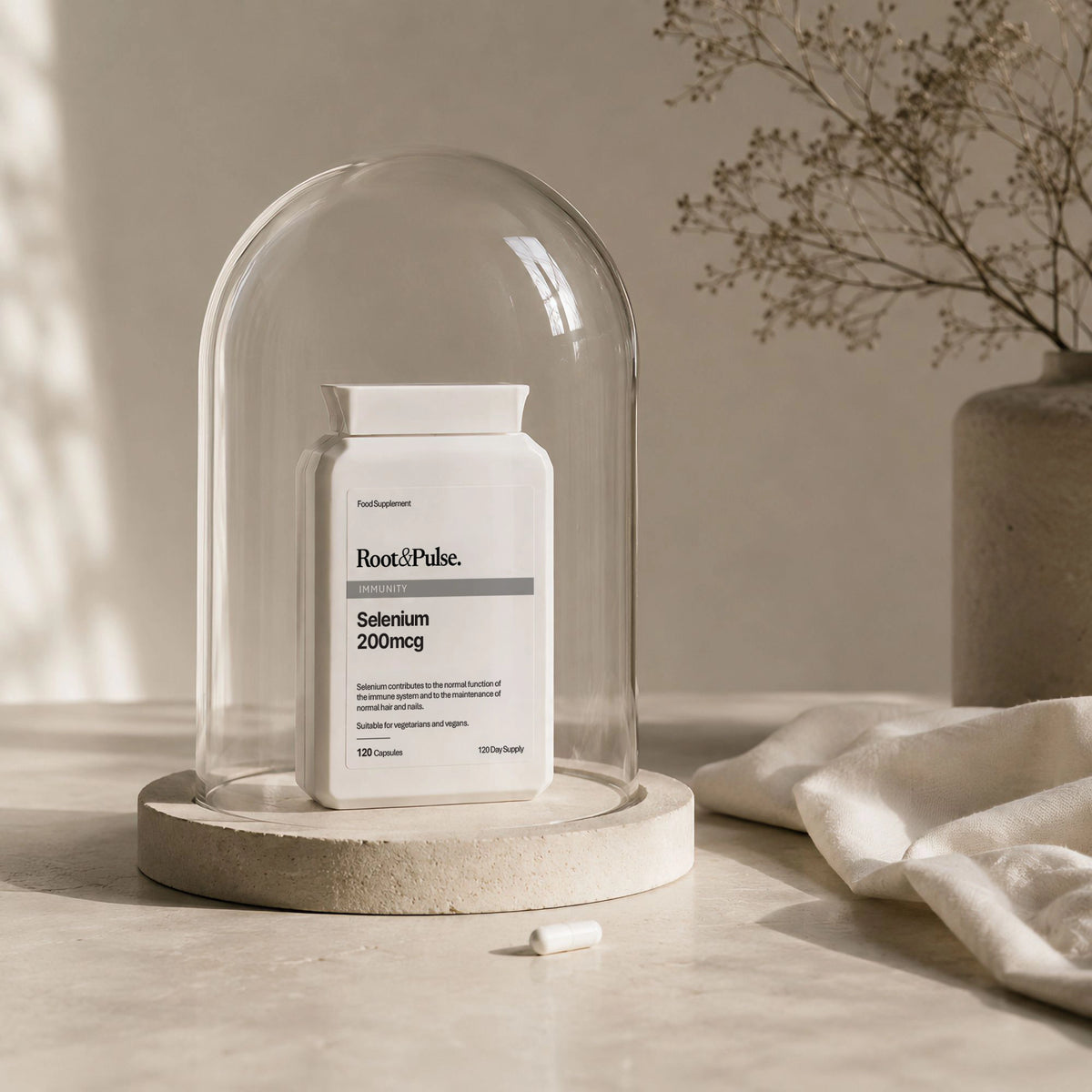 A white supplement bottle labeled Root and Pulse Selenium 200mcg sits under a clear glass dome on a round stone base, styled on a light surface with soft shadows and dried flowers in the background.