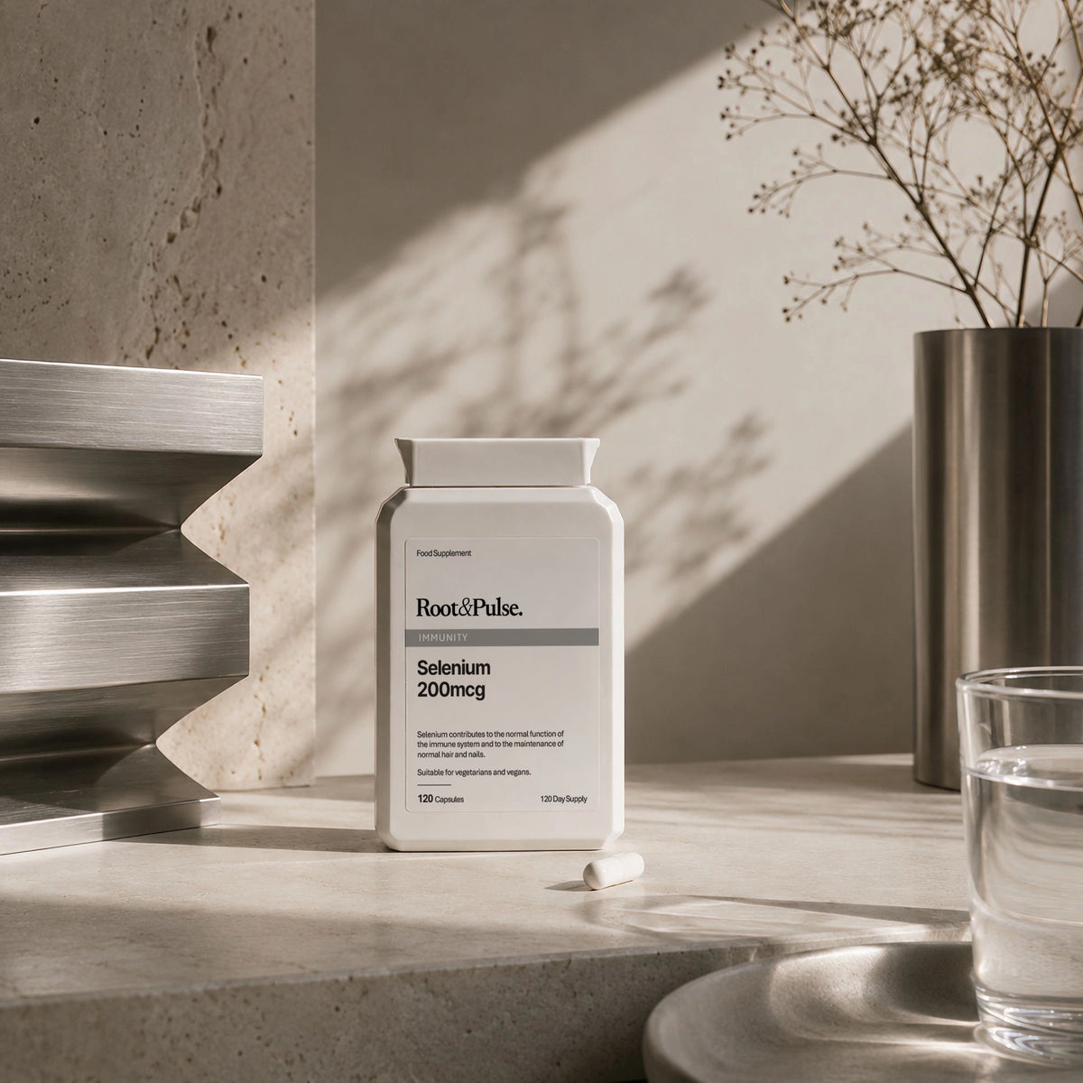 A white bottle of Root & Pulse Selenium 200mg capsules sits on a stone countertop in a modern, sunlit room with concrete walls, a glass of water, and dried branches in a vase.