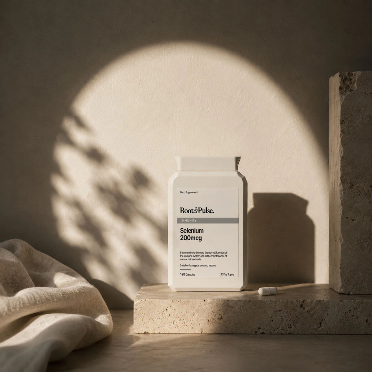 A white bottle of Root & Pulse. Selenium 200mg capsules sits on a textured stone pedestal with dramatic, warm sunlight casting a shadow on a beige wall.