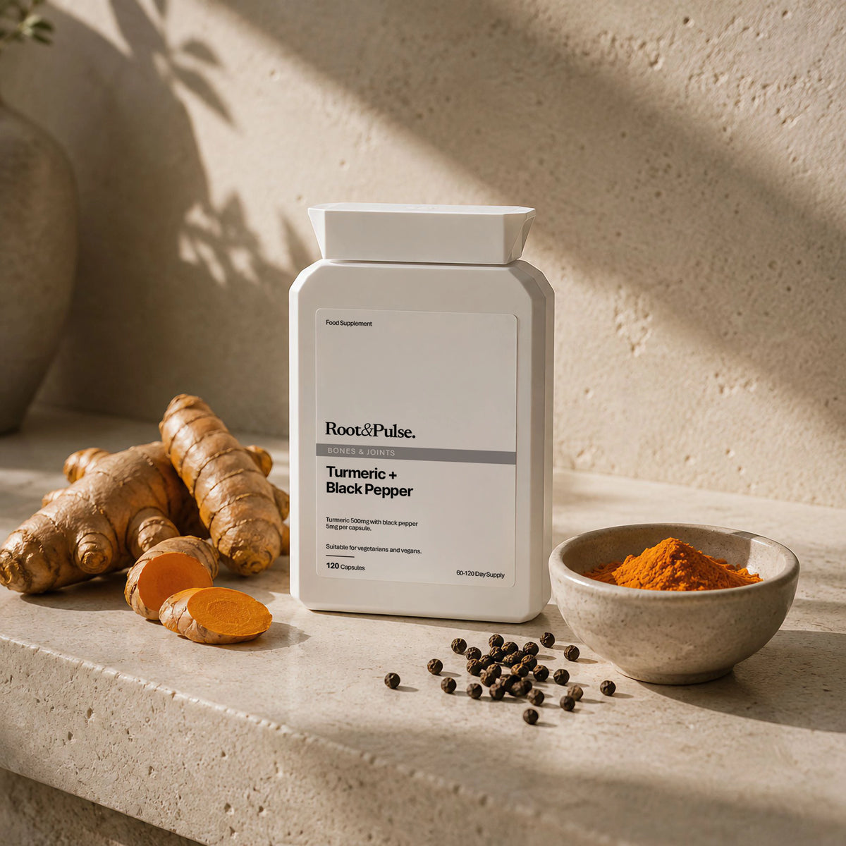 A white bottle of Root & Pulse Turmeric + Black Pepper supplement stands on a stone surface with turmeric roots, a bowl of orange powder, and scattered black peppercorns in warm natural light.
