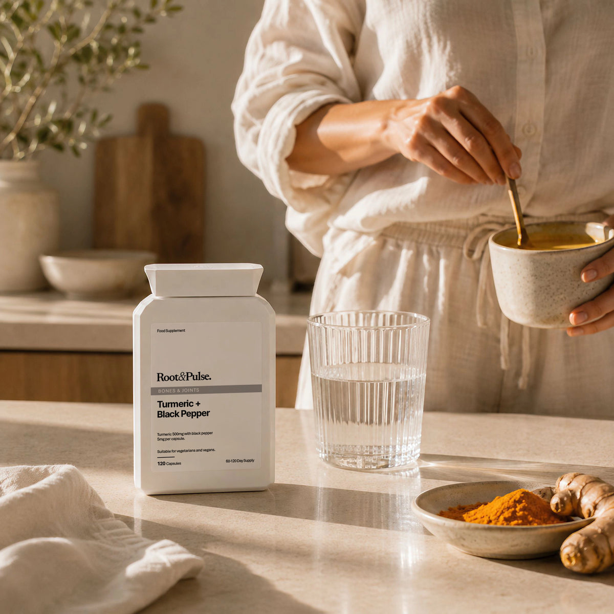 A person in a white robe stands in a warm kitchen setting, holding a bowl and spoon while preparing a drink. In the foreground on a light stone countertop sits a white bottle labeled Root and Pulse Turmeric + Black Pepper, alongside a glass of water, a small dish of orange powder, and turmeric roots.