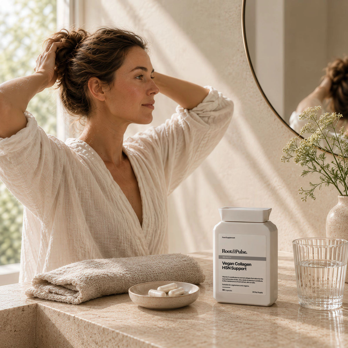 A woman in a white robe stands at a bathroom counter adjusting her hair, with a bottle of Root & Pulse Vegan Collagen Hair Support, a folded towel, a small dish of capsules, and a glass of water nearby.