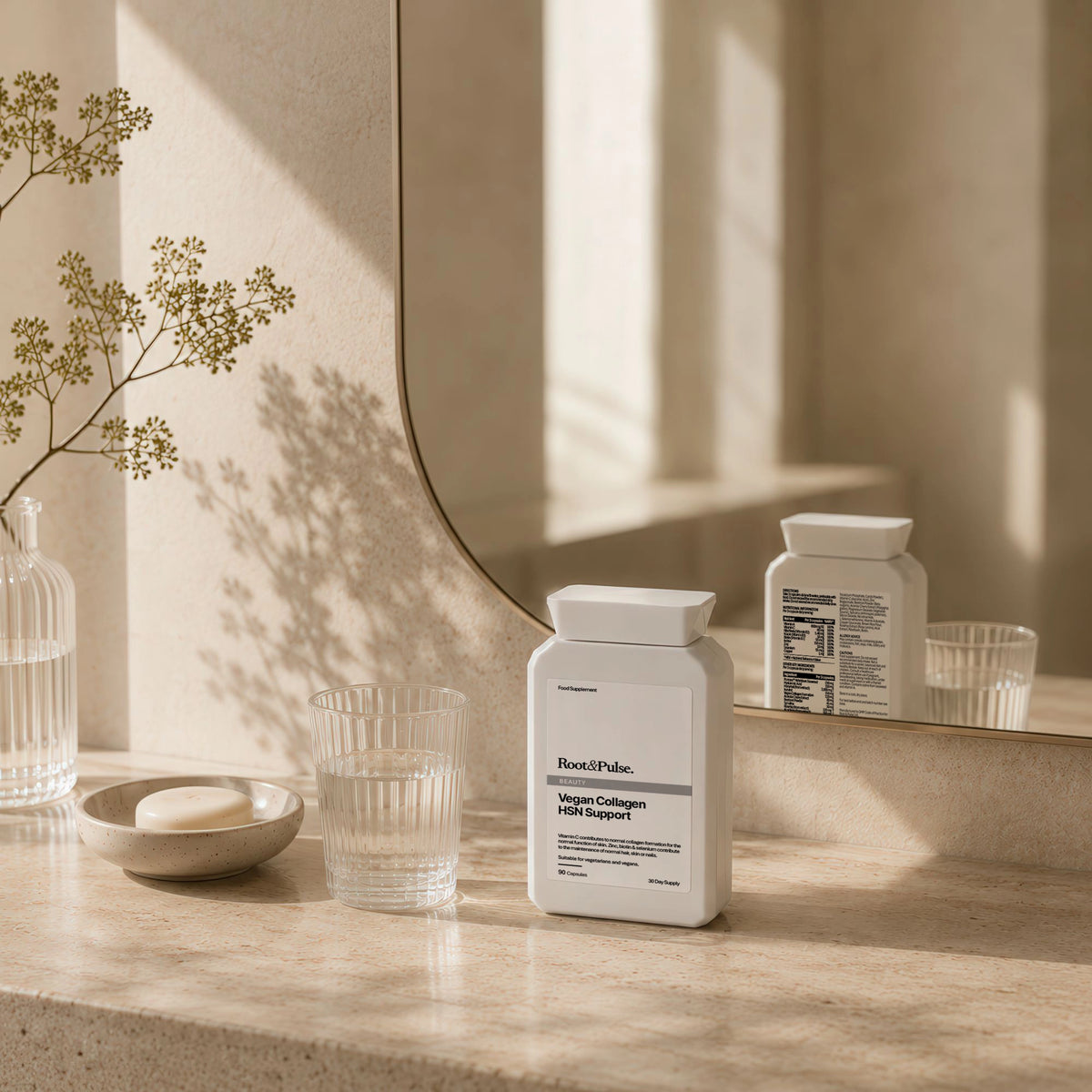 A white supplement bottle labeled Root & Pulse Collagen Supplement sits on a beige stone countertop beside a glass of water, a small dish with soap, and a mirror reflecting the bottle in warm natural light.