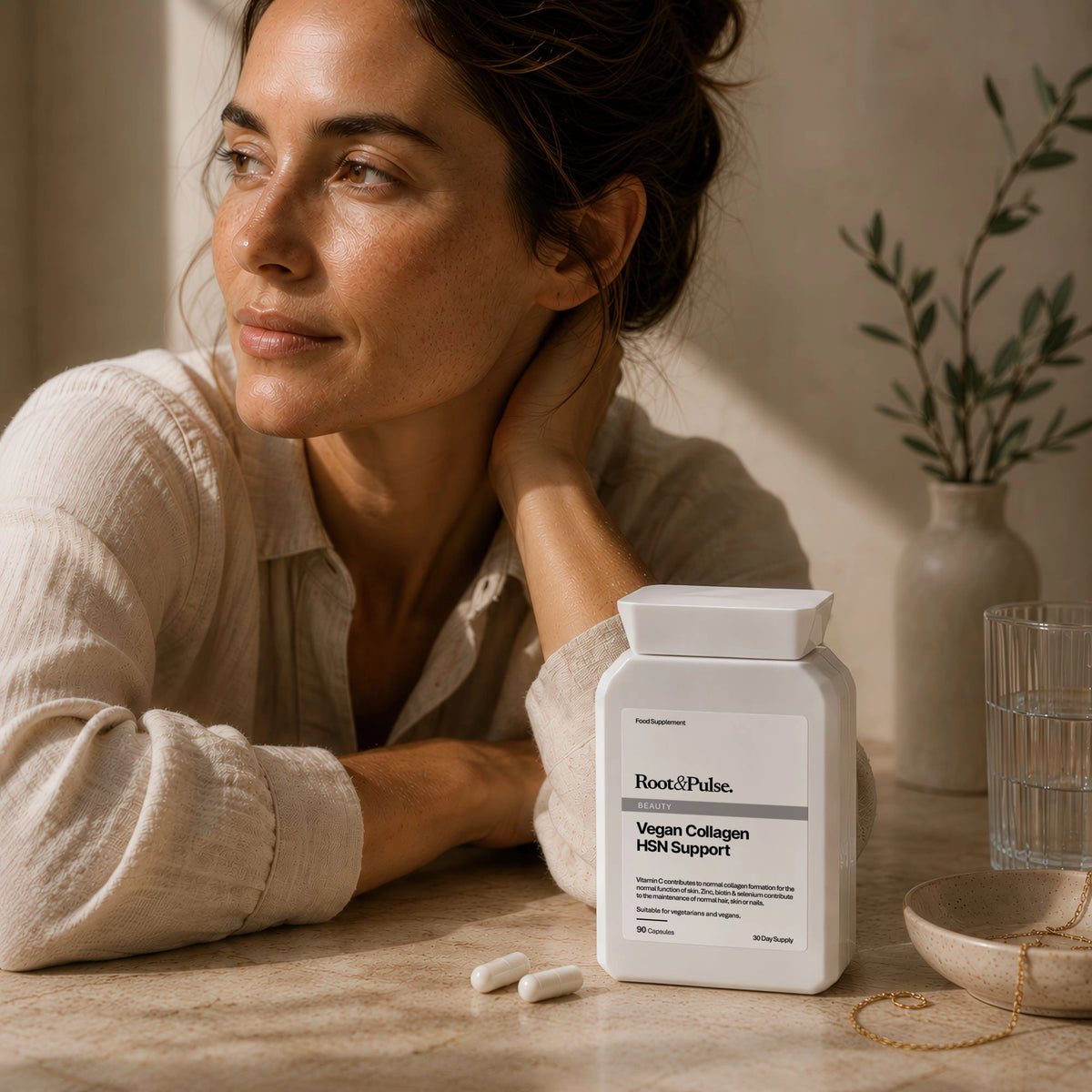A woman in a beige linen shirt rests her head on a stone table next to a white bottle of Root & Pulse Vegan Collagen Supplement and HSN, with two white capsules lying nearby and a glass of water in the background.