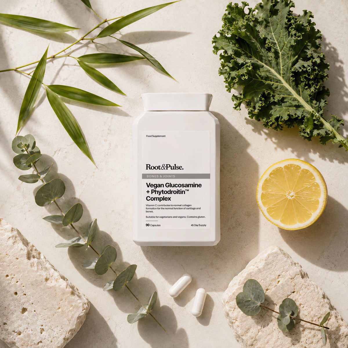 A white bottle of Root and Pulse Vegan Glucosamine + Phytodrin Complex dietary supplement, styled in a flat lay with green kale, a sliced lemon, eucalyptus leaves, and textured stone slices on a light beige background.