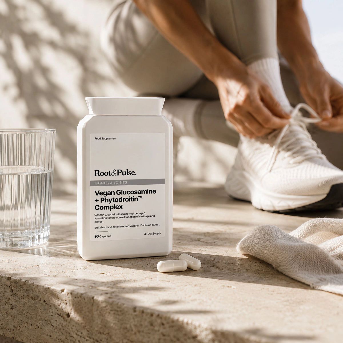 A white bottle of Root & Pulse Vegan Glucosamine + Phytodrotin Complex supplement sits on a stone ledge next to a glass of water, with a person sitting in the background tying white sneakers.