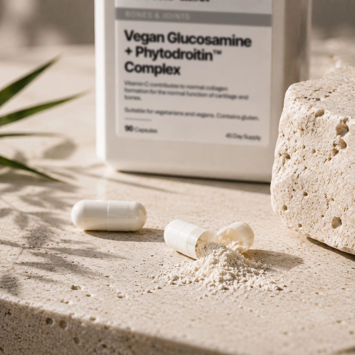 A white bottle labeled  Root & Pulse 'Vegan Glucosamine + Phytodroitin Complex' sits on a sunlit stone surface with two capsules and a small pile of powder in front. The scene includes natural shadows and a textured stone prop.