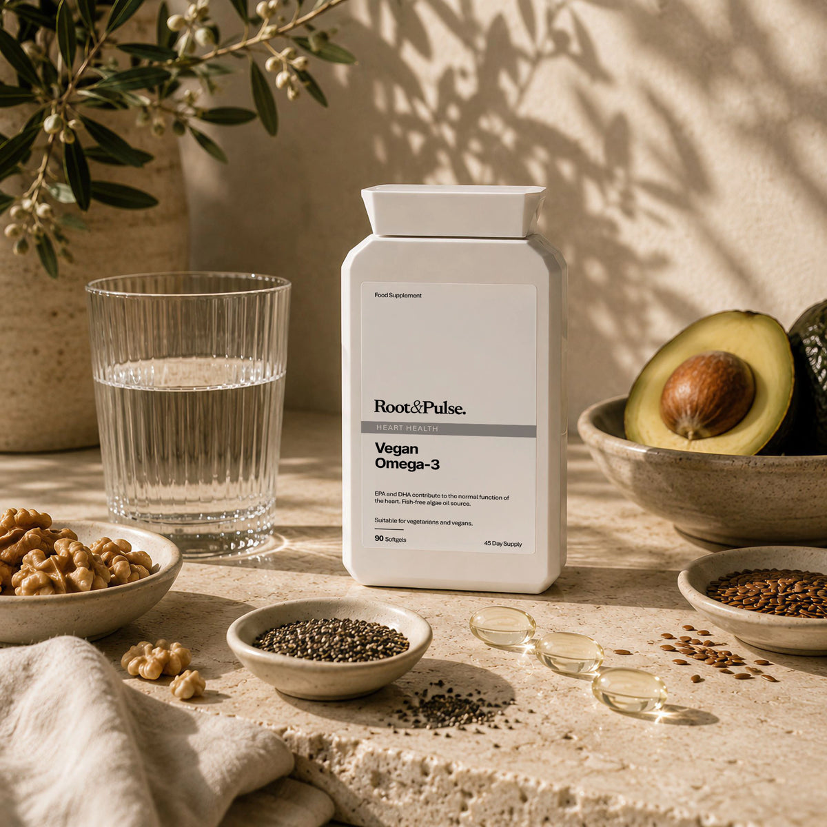 A bottle of Root&Pulse Vegan Omega-3 dietary supplements stands on a stone surface surrounded by natural ingredients including walnuts, chia seeds, flax seeds, a glass of water, and avocado, under warm natural sunlight with leaf shadows.