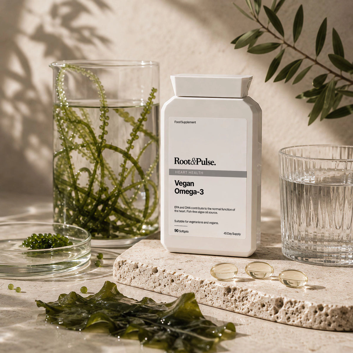 A bottle of Root & Pulse Vegan Omega-3 supplement stands on a stone slab beside a glass of water, with seaweed and capsules arranged around it in a styled studio setup.