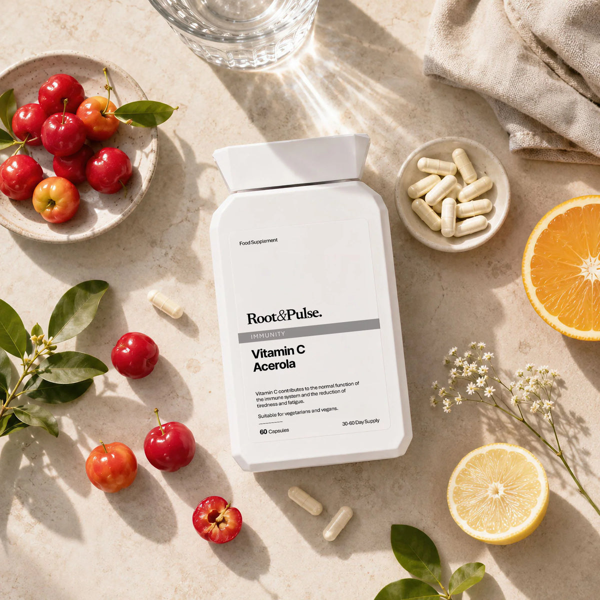 A white bottle of Root&Pulse Vitamin C Acrola dietary supplement capsules, styled in a flat-lay with fresh acerola cherries, an orange slice, a lemon slice, and small bowls of capsules on a light beige stone surface.