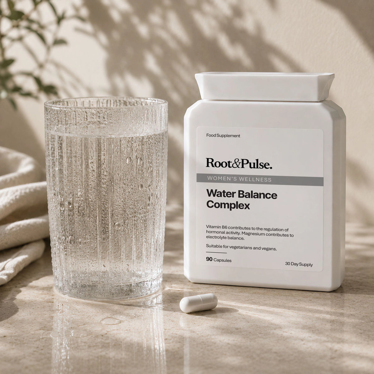 A white bottle of Root&Pulse Women's Wellness Water Balance Complex food supplement stands on a stone surface beside a textured glass of water and a single capsule, with soft natural shadows and a neutral fabric in the background.