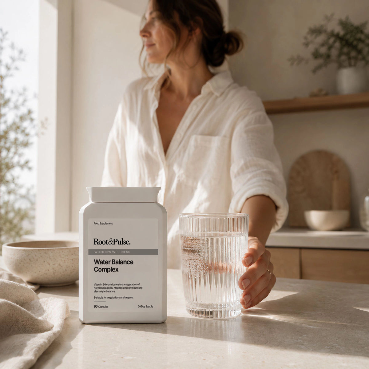 A woman in a white shirt stands in a bright kitchen holding a glass of water next to a white bottle of Root and Pulse Water Balance Complex.