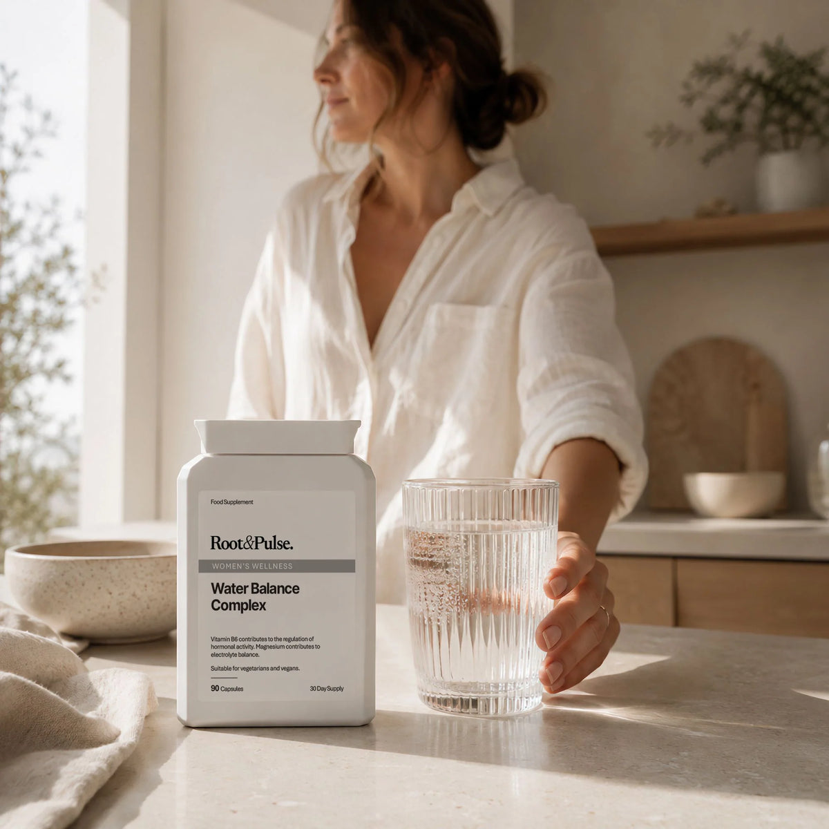 A woman in a white shirt stands in a bright kitchen holding a glass of water next to a white bottle of Root and Pulse Water Balance Complex.