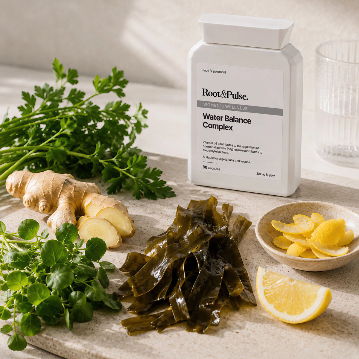 A bottle of Root & Pulse Water Balance Complex dietary supplement stands on a stone surface with its ingredients arranged around it: fresh ginger, green herbs, seaweed, lemon slices, and a glass in a bright, clean setting.