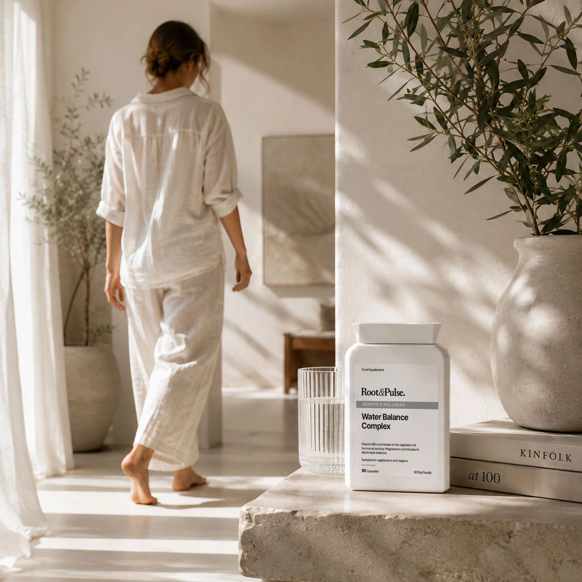 A woman in white pajamas walks barefoot through a sunlit room, while a white Root & Pulse Water Balance Complex supplement bottle sits on a stone ledge in the foreground with a glass and books nearby.