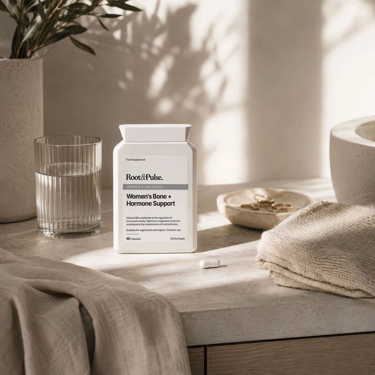 A white supplement bottle labeled Root of Pulse Women's Bone + Hormone Support sits on a stone countertop in a softly lit bathroom setting with a glass of water, folded towel, and small dish nearby.
