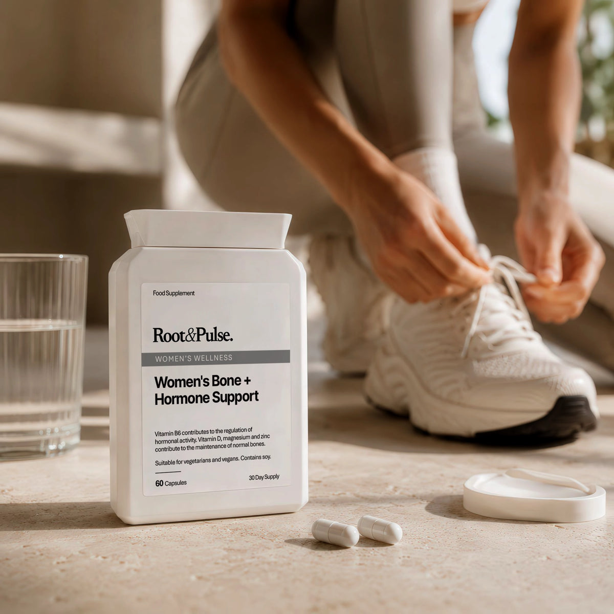 A bottle of Root & Pulse Women's Bone + Hormone Support dietary supplement stands on a stone surface next to a glass of water, with a person in the background tying their white sneakers.