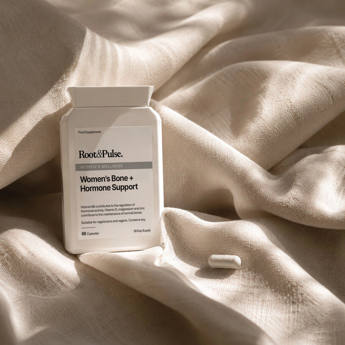 A white supplement bottle labeled Root&Pulse Women's Bone + Hormone Support sits on draped cream fabric with a single capsule beside it.