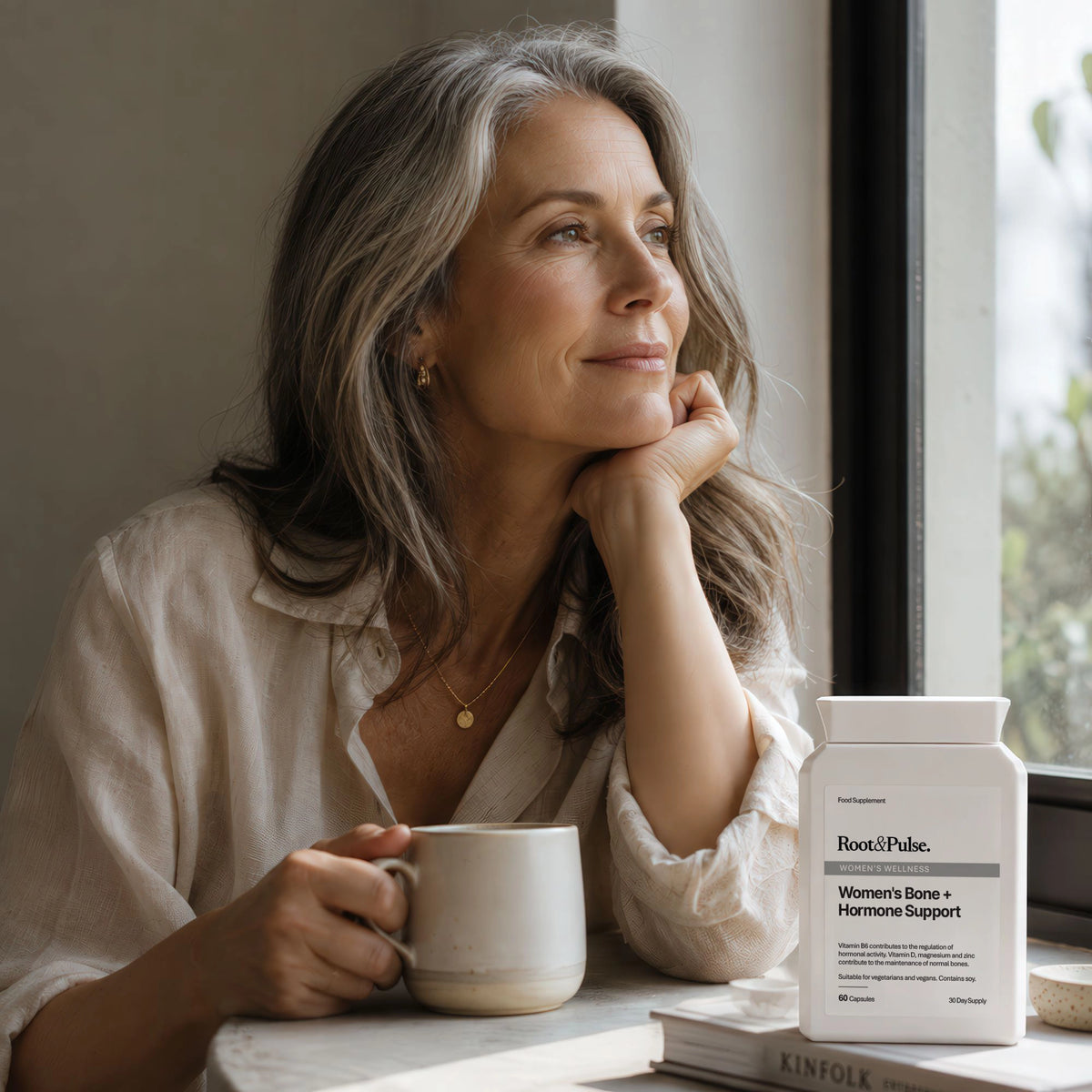 A woman with gray hair sits by a window in a bright white shirt, holding a ceramic mug. A white supplement bottle labeled Root of Paliu Women's Bone + Hormone Support sits on the table beside her.