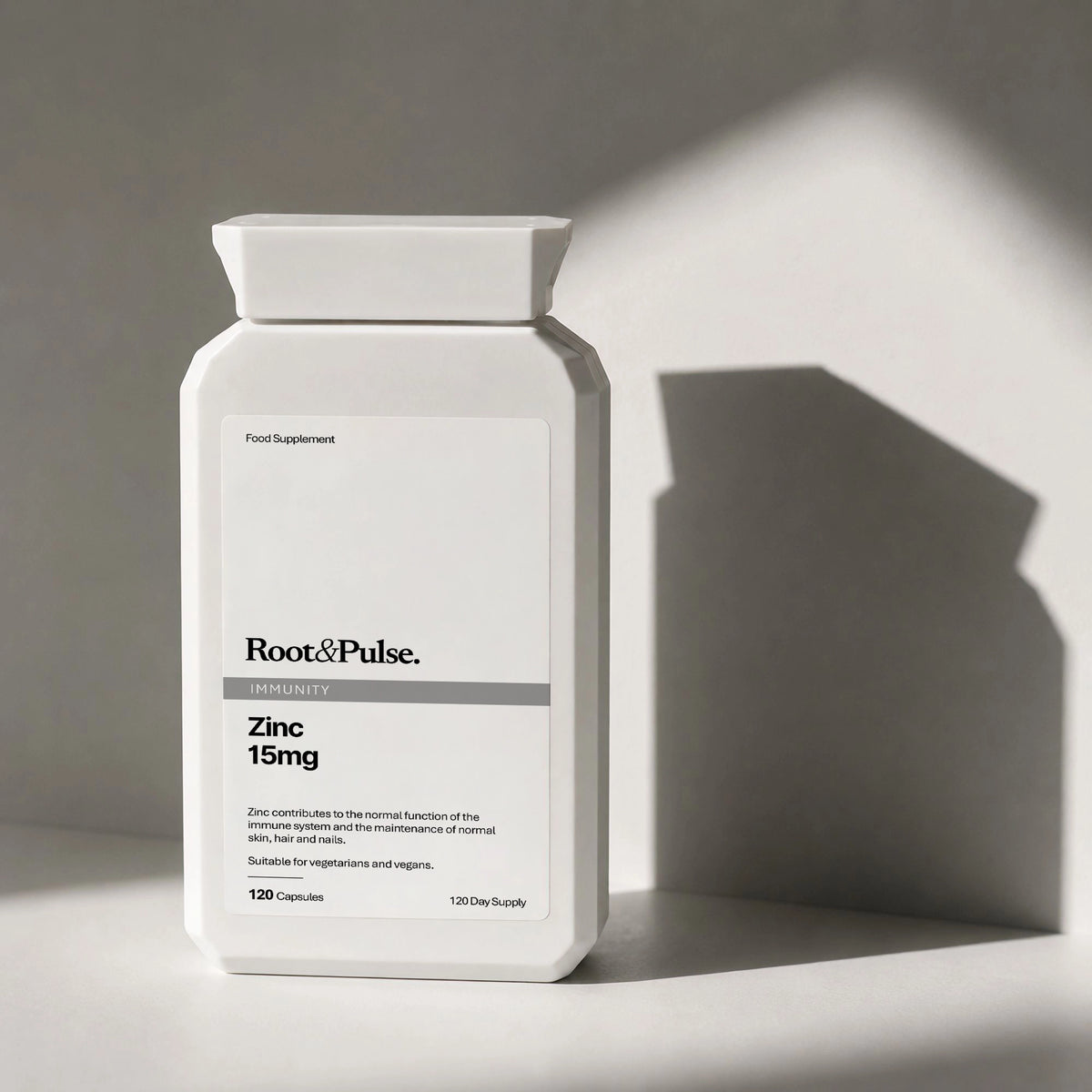 A white plastic bottle of Root & Pulse Zinc 15mg food supplement, photographed on a gray surface with dramatic diagonal shadows.