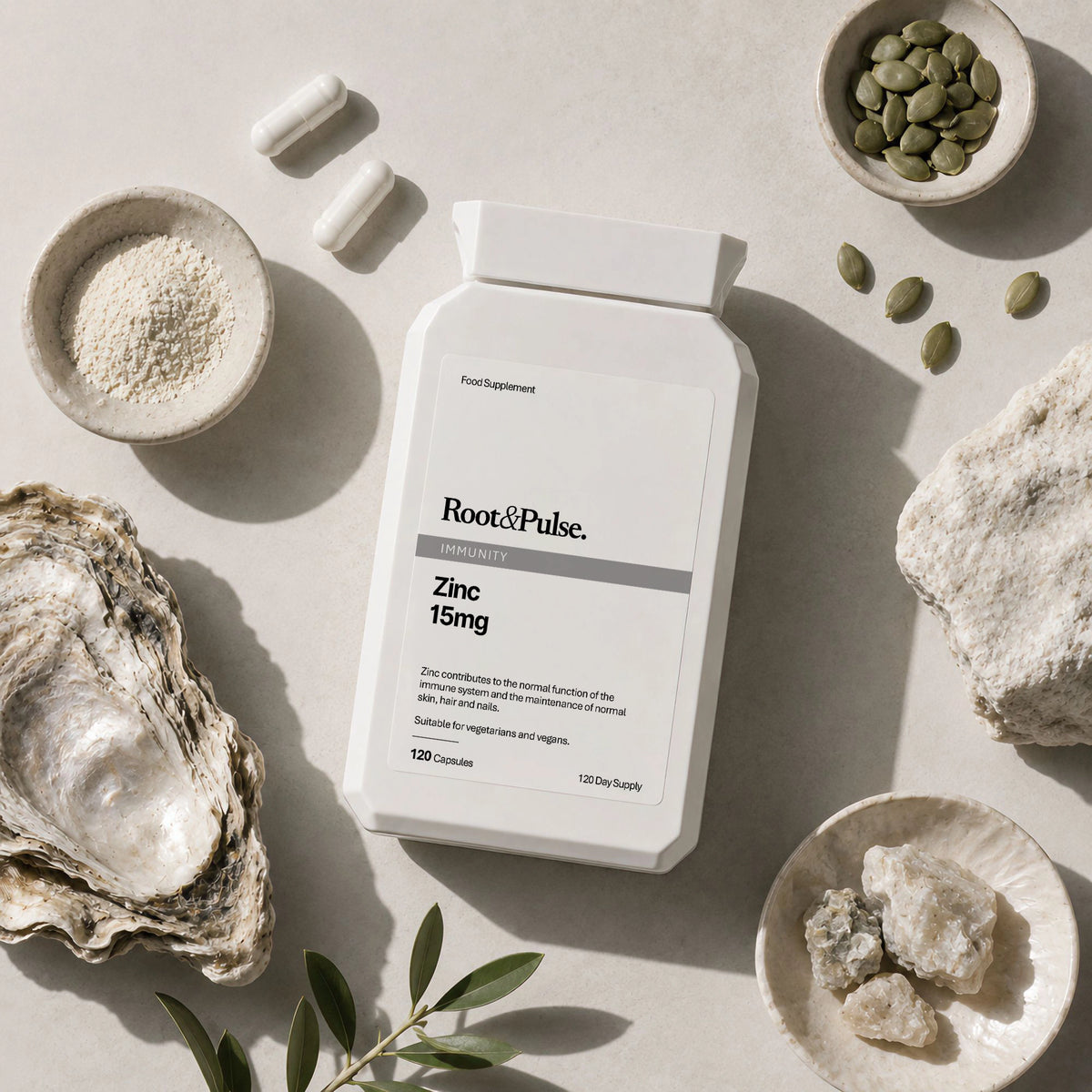 A white supplement bottle labeled Root of Pulse Zinc 15mg, styled in a flat-lay with an oyster shell, green seeds, and small bowls of ingredients on a light stone surface.