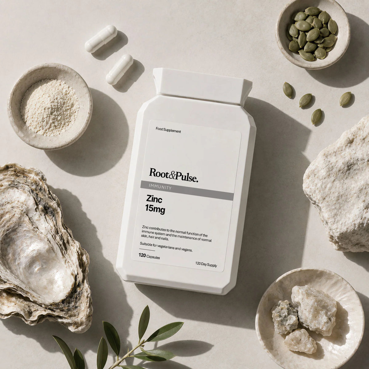 A white supplement bottle labeled Root of Pulse Zinc 15mg, styled in a flat-lay with an oyster shell, green seeds, and small bowls of ingredients on a light stone surface.