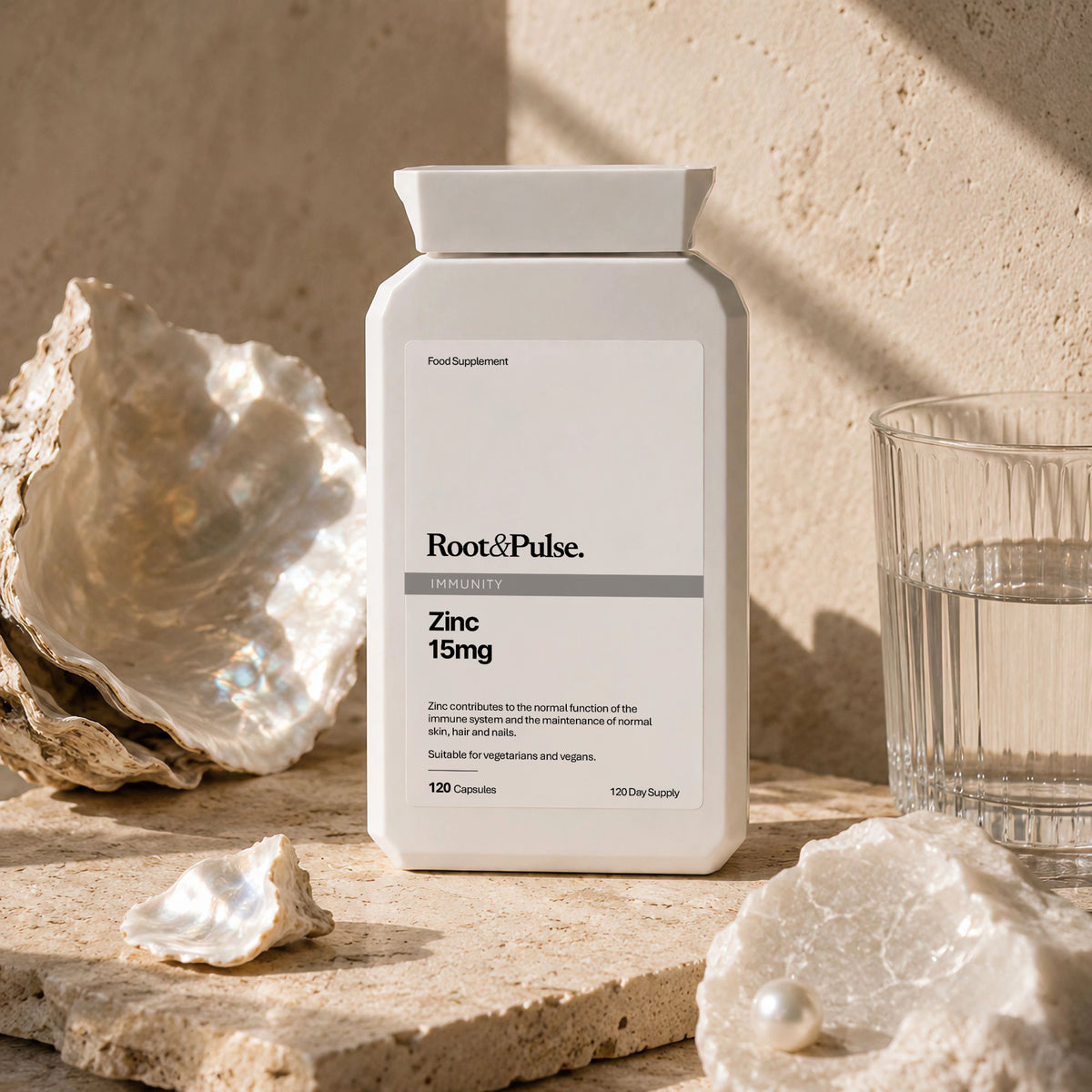 A white bottle of Root & Pulse Zinc 15mg dietary supplement, styled on a beige stone surface with oyster shells and a glass of water.