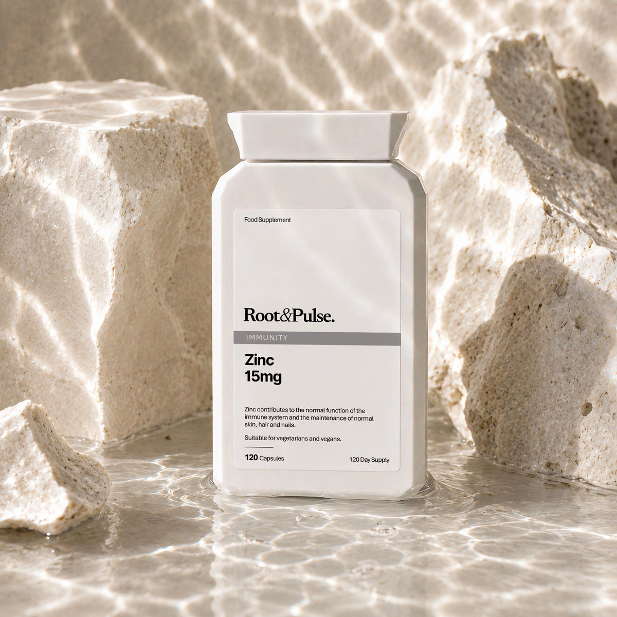 A bottle of Root & Pulse Zinc 15mg dietary supplement, styled on a sandy beach with water ripples and white rocks in the background.